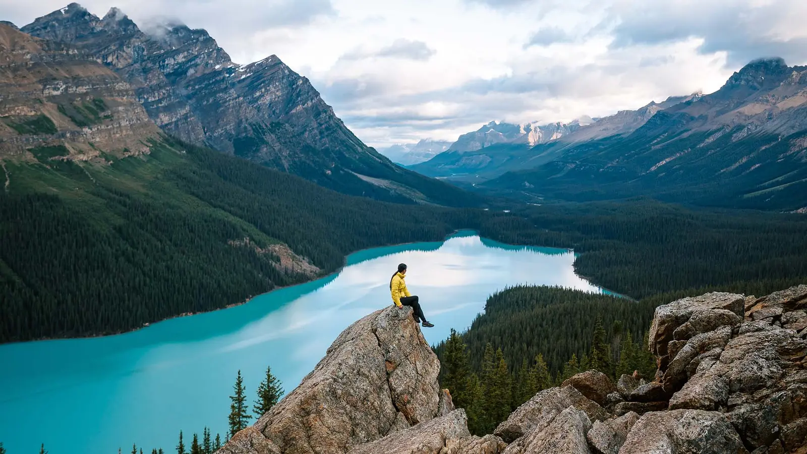 A traveler sits on a rock above a turquoise mountain lake surrounded by forest
